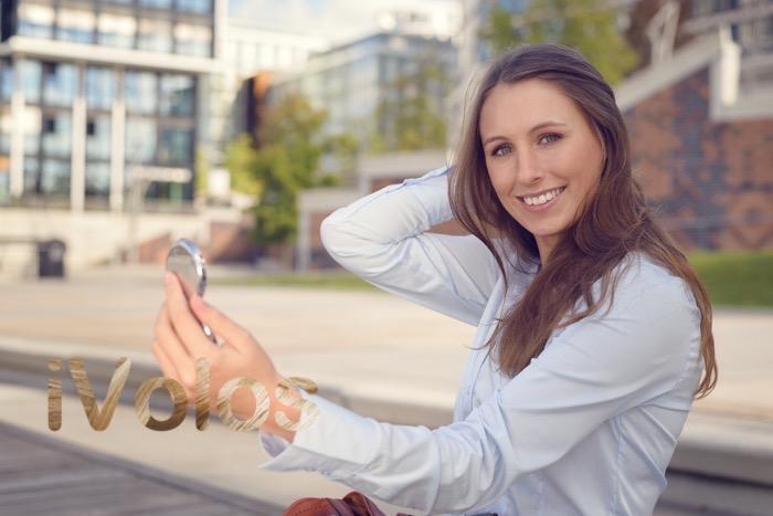 Beautiful young woman checking her appearance in a small handheld mirror in the street and smiling as she holds her long brown hair away from her face